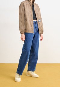 Hellbraune, taillierte Bomberjacke aus strukturiertem Stoff, mit gerippten Bündchen und seitlichen Taschen, kombiniert mit hochtaillierten blauen Jeans und beigefarbenen Turnschuhen.