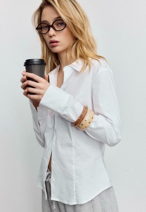 Camicia bianca con bottoni e maniche lunghe, indossata con pantaloni grigi. Accessoriata con braccialetti di legno e una tazza di caffè nera. Tessuto liscio, vestibilità rilassata.