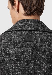 Grå struktureret frakke med stor krave, prydet med en sort læderlabel på ryggen med "ALLSAINTS" og en vævet stofstruktur.