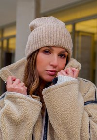 Beige knitted beanie and oversized cream shearling jacket with black accents, featuring a soft texture and relaxed fit.