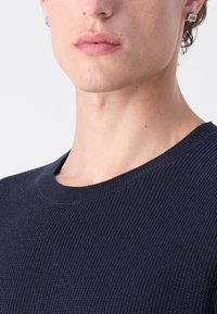 Maglione in punto navale, lavorato a maglia con scollo rotondo. Vista ravvicinata mostra i dettagli del tessuto e del colletto. Orecchino argentato visibile.