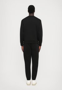 Man standing facing away wearing a black sweatshirt and sweatpants with white sneakers against a plain white background.