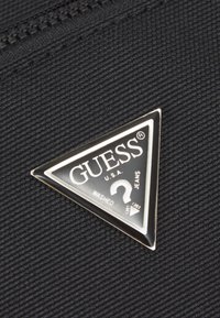 Tissu noir texturé avec un logo triangulaire argenté comportant "GUESS U.S.A." et les mots "WASHED JEANS" en texte blanc.