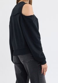 Femme portant un haut noir à épaules dénudées à manches longues et un jean gris foncé, debout de profil sur un fond clair uni.