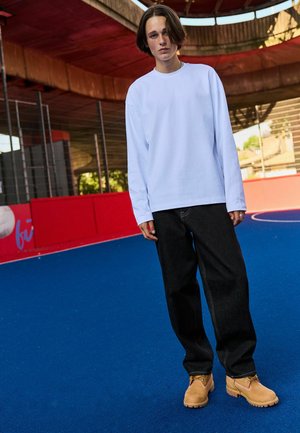 Long-sleeve white t-shirt paired with dark wash baggy jeans and tan suede boots, standing on blue synthetic field with red accents.