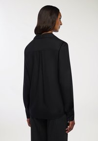 Czarna, długozasłonięta koszula z gładkiego materiału, z tylnym karczkiem, mankietami na guziki i zaokrąglonym dołem. Noszona z spodniami w paski.