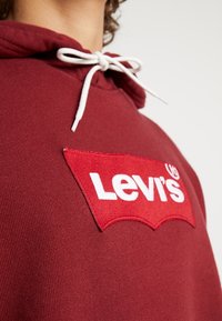 Felpa cappuccio bordeaux con logo Levi's ricamato in rosso sul petto, con cordini bianchi attaccati al cappuccio. Tessuto morbido e lavorato.