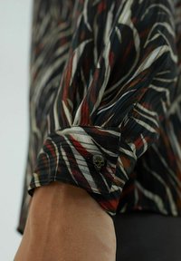 Blusa nera con stampe astratte in rosso e bianco; presenta un bottone a forma di teschio sul polsino; tessuto leggero e plissettato con un design fluido.