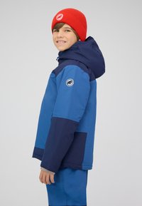 Ragazzo che indossa una giacca blu e blu scuro con cappuccio e pantaloni blu, sorridente, e un berretto di lana rosso con un logo circolare bianco.