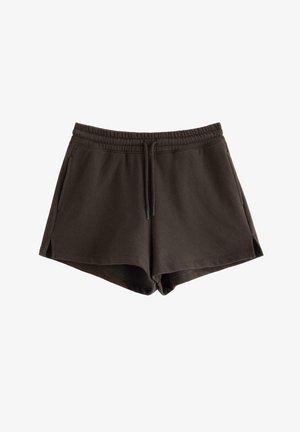 Kvinde står barfodet med den ene hånd på hoften iført en lys pink crop top og mørke løse shorts mod en ensfarvet lys baggrund.