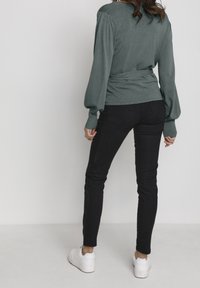 Tmavě zelený svetr s dlouhými rukávy a nabíranými rameny, s detailem na zavázání v pase, spárovaný s černými skinny džíny a bílými botami. Minimální textura.