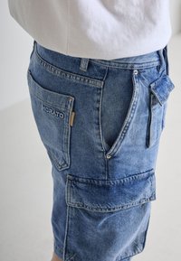 Denim cargo kratke hlače u svijetloplavoj boji, s višestrukim džepovima, dekorativnim šavovima i oznakom brenda. Tkanina ima teksturiranu završnu obradu.