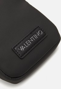 Custodia per telefono nera con texture, dotata di un'etichetta logo in rilievo con la scritta "VALENTINO". Finitura liscia con un sottile motivo a righe lungo la superficie.