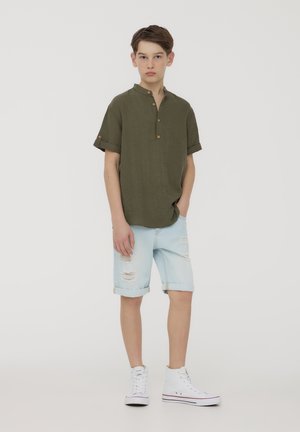 Camicia corta verde oliva con colletto e bottoni marroni, abbinata a pantaloni shorts in denim chiaro strappati e sneakers bianche alte.