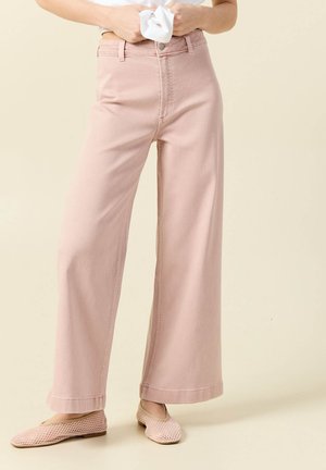 Mujer con pantalones de tiro alto y perneras anchas de color rosa claro y zapatos planos texturizados a juego, sujetando un paño blanco en la cintura.