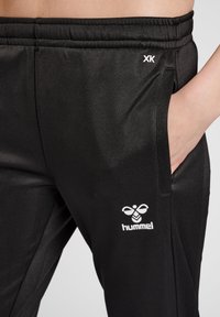 Schwarze Sporthosen mit glatter Textur, ausgestattet mit einem weißen Hummel-Logo und einer Tasche. Elastischer Bund für zusätzlichen Komfort.