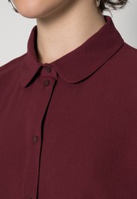 Bordeaux blouse met een kraag, textuurstof, vier knoopsluiting en subtiele contrasterende stiknaad op de sluiting.