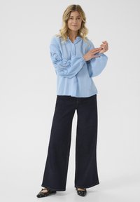 Blusa azzurro chiaro con bottoni e maniche a palloncino ricamate, abbinata a jeans svasati scuri e scarpe nere flat con borchie decorative.