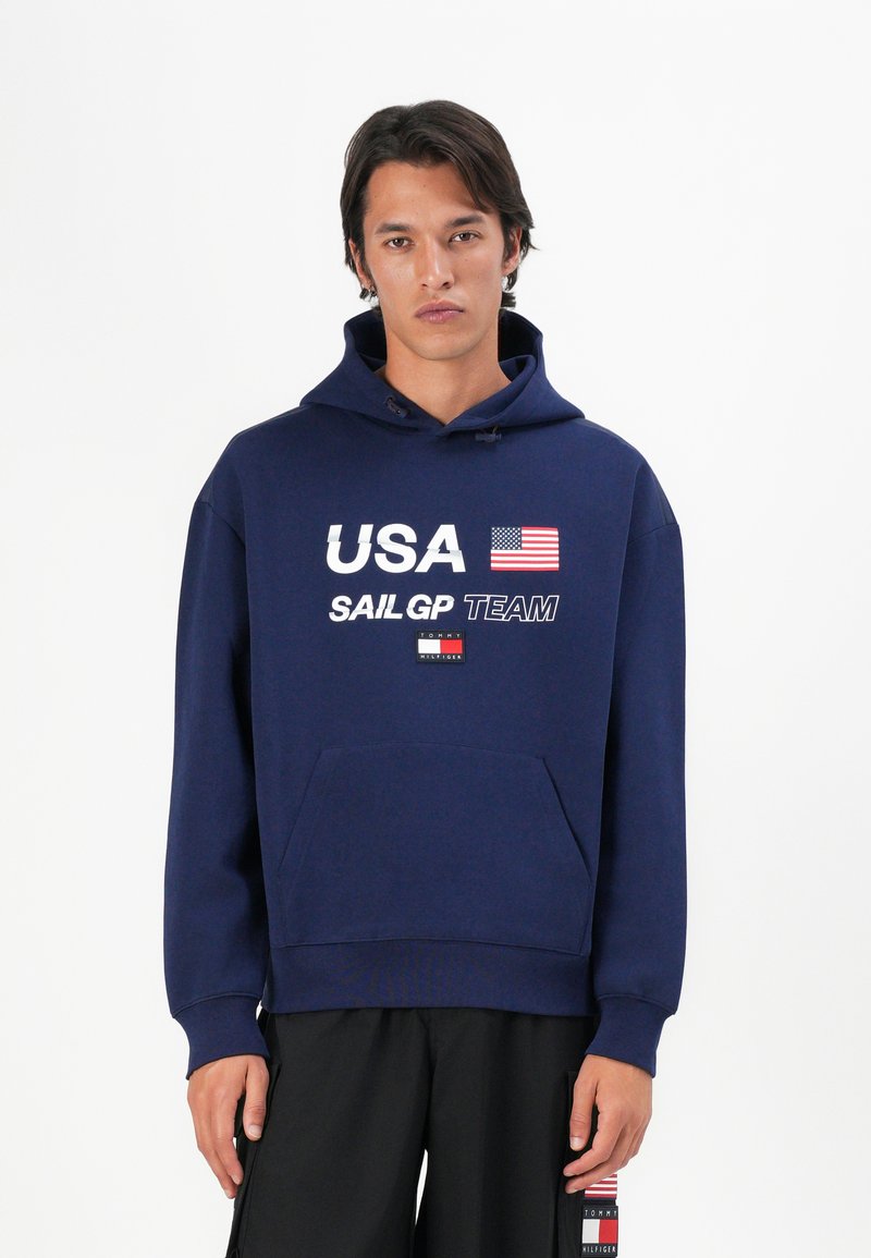 Felpa con cappuccio blu navy con la scritta "USA SAILGP TEAM", grafica della bandiera americana, tasca frontale e cappuccio regolabile con cordino. Materiale in morbido misto cotone.