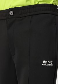 Juodos kelnės, pagamintos iš lygios, tamprios medžiagos, su mygtuku ir siuvinėtu logotipu, kuriame parašyta "nauji originalai", šone.