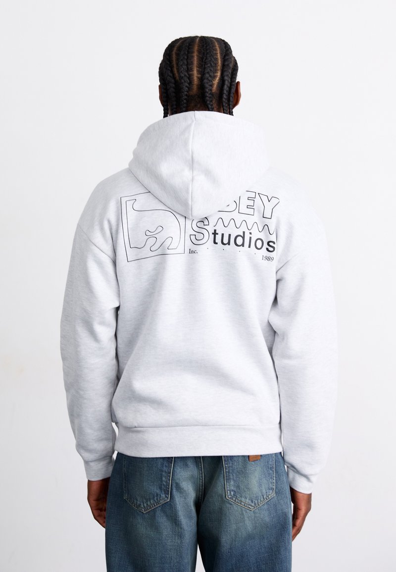 Light gray hoodie with a hood, featuring a black graphic logo on the back, and ribbed cuffs and hem. Paired with blue denim jeans.