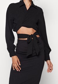 Blouse noire à manches longues en tissu texturé. Ceinturée à la taille avec une ceinture assortie, associée à une jupe noire moulante.
