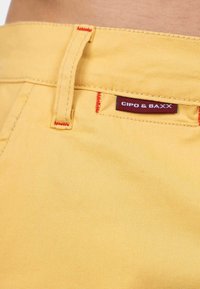 Pantaloni in tessuto giallo con una trama liscia, dotati di un'etichetta e cuciture rosse a contrasto sulla vita e sulle tasche.