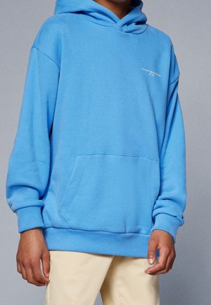 Niebieska bawełniana bluza z kapturem z kieszenią z przodu, ściągaczami na mankietach i u dołu. Na piersi znajduje się biały nadruk z logo, a kaptur ma sznurek do regulacji.