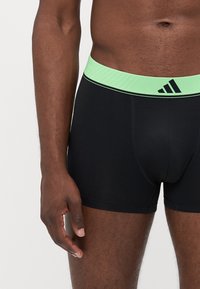 Črne bokserice z elastičnim pasom v zeleni barvi, na katerem je logo Adidas. Tkanina ima gladko teksturo in prilega se telesu.