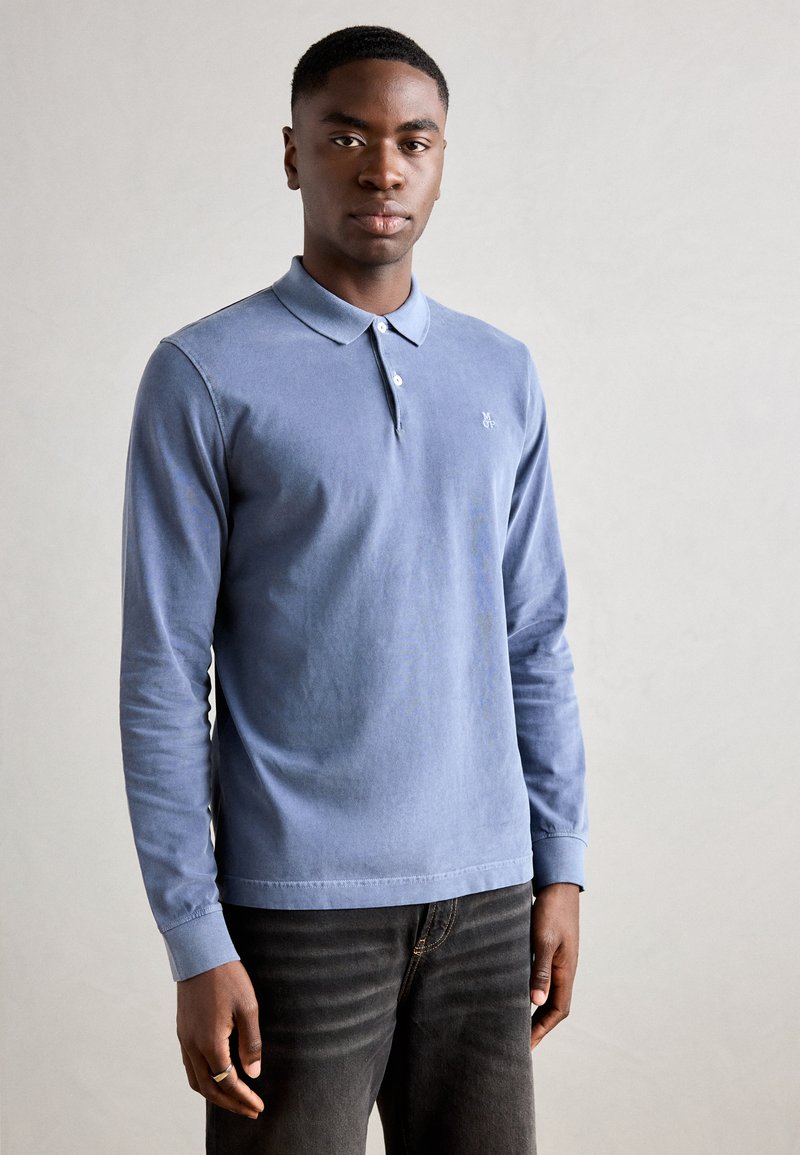 Polo shirt bleu à manches longues avec col côtelé, patte de boutonnage à deux boutons et logo sur la poitrine, associé à un jean foncé.