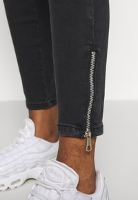 Tobillo vistiendo pantalones negros con detalles de cremallera plateada y zapatillas Air Max blancas sobre un fondo claro.