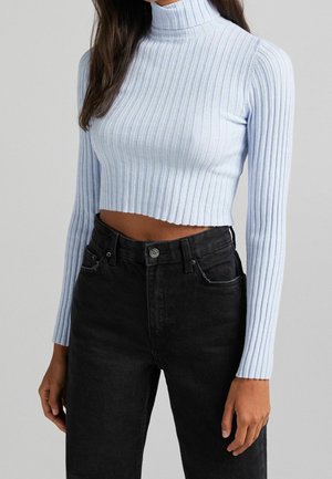 Nainen pukeutuneena vaaleansiniseen ribbineulottuun poolokaulukselliseen lyhytmalliseen neulepaitaan ja korkeavyötäröisiin mustiin farkkuihin, yksiväristä harmaata taustaa vasten.