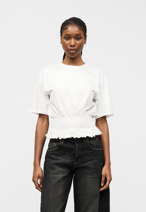 Junge Frau mit geflochtenem Haar, die ein weißes kurzärmeliges Oberteil mit elastischem Bund und schwarze High-Waist-Jeans trägt, vor einem schlichten Hintergrund steht.