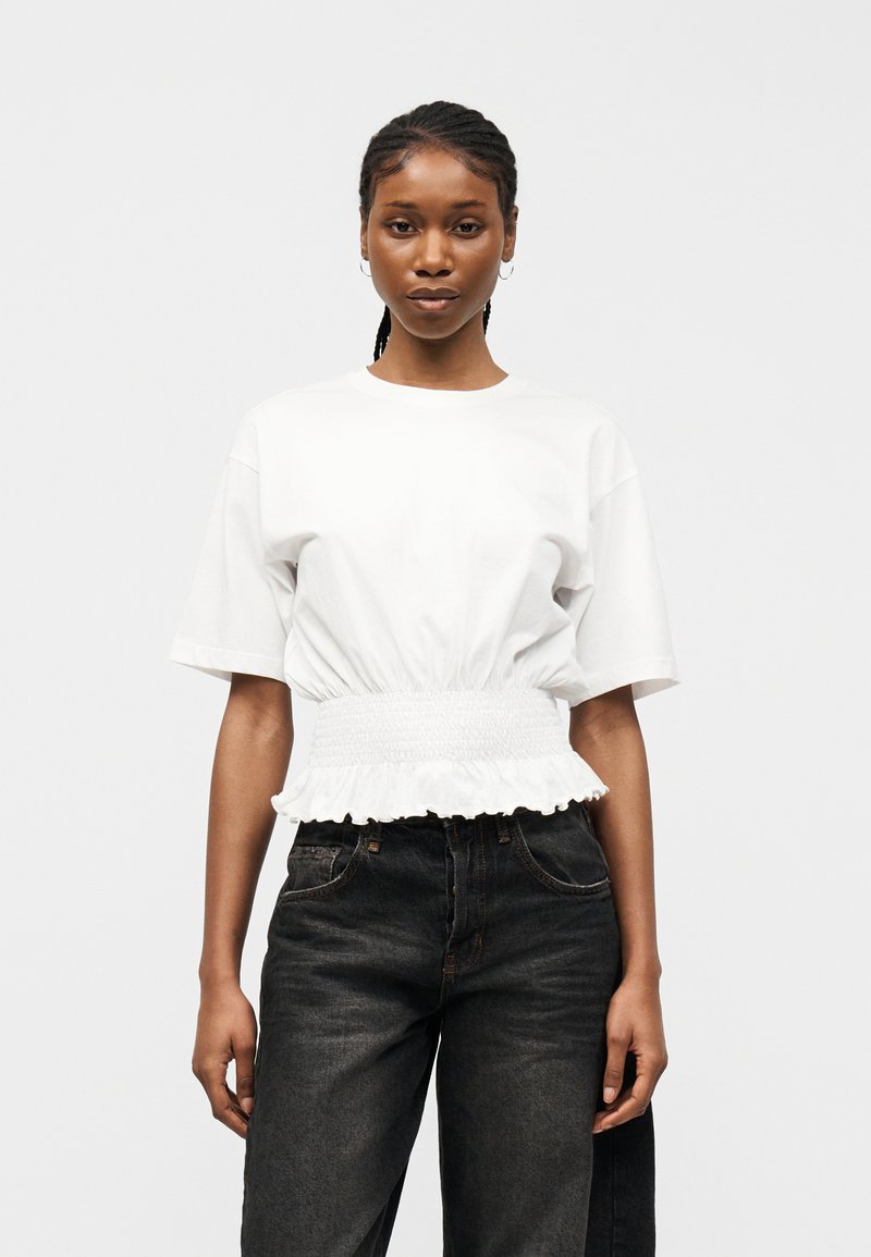 Νεαρή γυναίκα με πλεγμένα μαλλιά, ντυμένη με λευκό κοντομάνικο τοπ με ελαστική μέση και μαύρα τζιν ψηλόμεσα, στέκεται μπροστά σε ένα απλό φόντο.