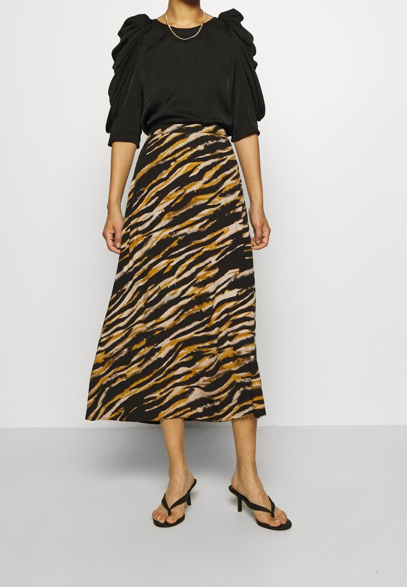 Femme portant un haut noir à manches bouffantes, un collier chaîne doré, une jupe midi imprimée tigre et des sandales à talons noires, debout sur un sol gris.
