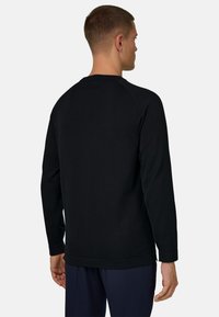 Homme de dos, portant un sweat-shirt à manches longues noir uni et un pantalon foncé, sur un fond gris clair.
