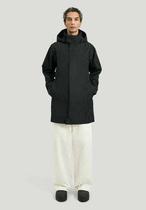 Person trägt schwarzen Kapuzenmantel, weiße weit geschnittene Hosen und schwarze Schuhe und steht vor einem schlichten hellgrauen Hintergrund.