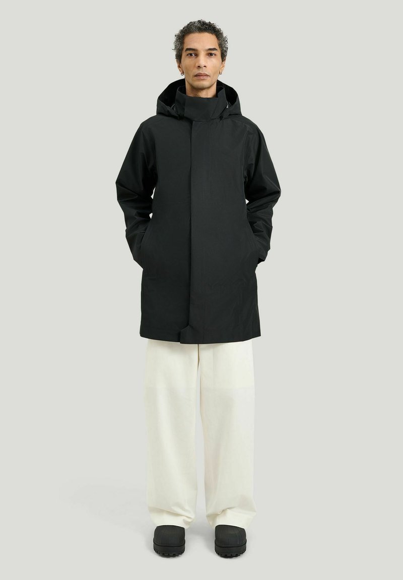 Person iført en sort hættejakke, hvide bukser med vide ben og sorte sko, stående mod en ensfarvet lys grå baggrund.