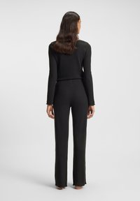 Γυναίκα που στέκεται ξυπόλητη, γυρισμένη προς τα πίσω, φορώντας εφαρμοστή μαύρη μπλούζα με μακρύ μανίκι και ίσια μαύρη παντελόνα με ελαφρώς ρυτιδιασμένες άκρες.