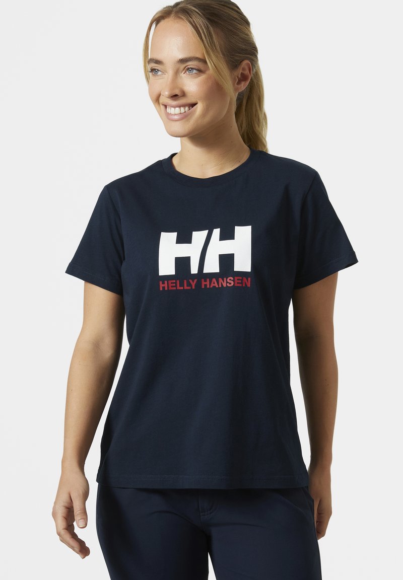 Mujer con el cabello rubio recogido, con una camiseta azul marino de Helly Hansen y pantalones a juego, sonriendo y mirando hacia un lado.