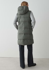 Padded Weste in Olivgrün mit Kapuze, horizontalen Nähten und kontrastierendem grauem Langarmoberteil. Trägt sie über dunklen Schlagjeans.
