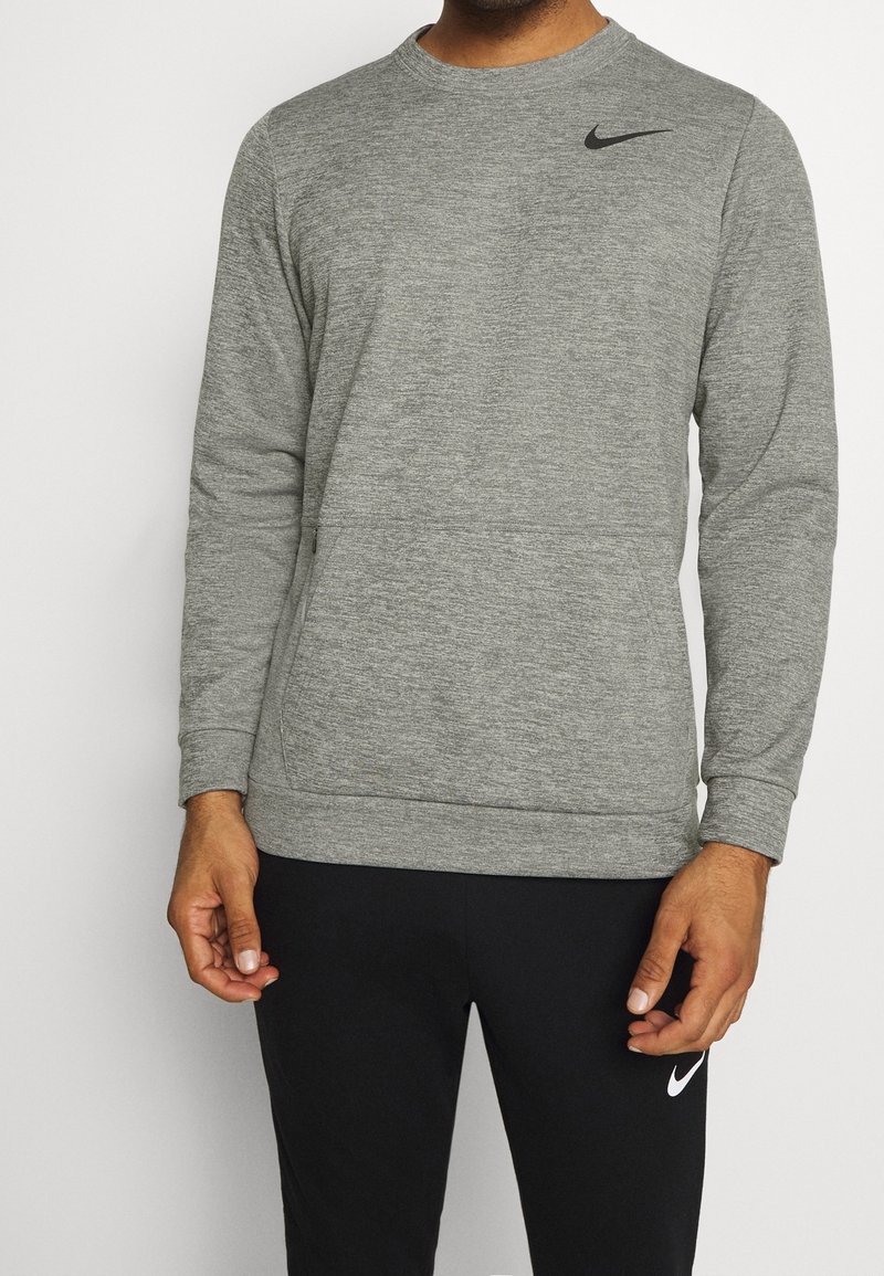 Homme portant un sweat-shirt Nike gris à manches longues et un pantalon Nike noir, debout les mains détendues le long du corps devant un fond blanc.