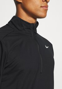 Maglia nera da atletica a mezza zip realizzata in tessuto testurizzato. Presenta un collo alto, un logo argentato sul petto e un design aderente per facilitare il movimento.