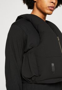 Colete acolchoado preto com colarinho alto, apresentando um fecho frontal com zíper, faixa elástica na cintura e um bolso plano no peito para armazenamento.