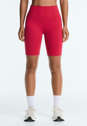 Predný pohľad na osobu, ktorá má na sebe jasne červené bicyklové krátke nohavice s vysokým pásom, biele ponožky a béžové tenisky na jednofarebnom pozadí.