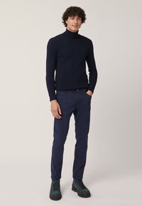 Maglione a collo alto a coste blu navy, pantaloni blu navy e stivali verde scuro. L'outfit presenta una vestibilità aderente con tessuto testurizzato e un discreto dettaglio del logo.