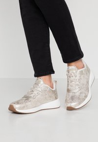 Zapatillas con una parte superior texturizada en dorado metálico, cordones grises y suela blanca acolchada, exhibidas con jeans negros sobre un fondo claro.