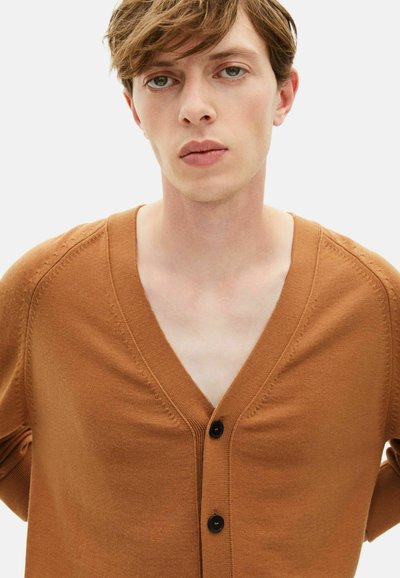Brauner Cardigan aus weichem Strickmaterial, mit einem tiefen V-Ausschnitt, vier schwarzen Knöpfen und gestrickten Details an den Schultern.