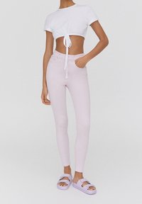 Crop top blanc avec détail noué, jean skinny rose pâle, et sandales lilas à double brides. Tissu lisse et coupe ajustée.