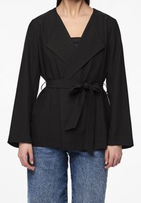 Veste enveloppante noire à manches larges, taille ceinturée et texture lisse. Présente un col en V profond et des poches à l'avant. Associée à un jean bleu.